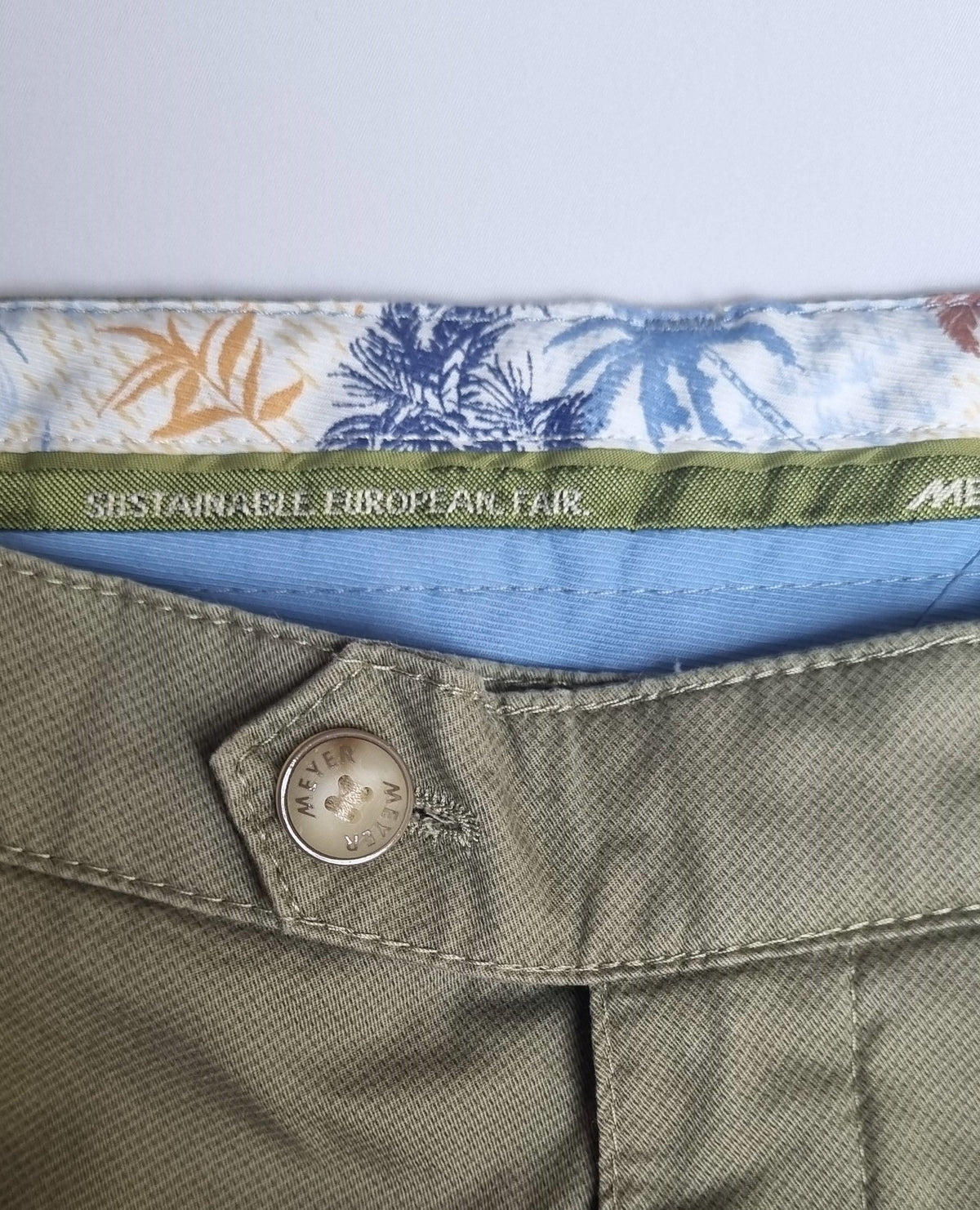 Close-up of a button on green pants with a floral patterned belt and visible brand name.